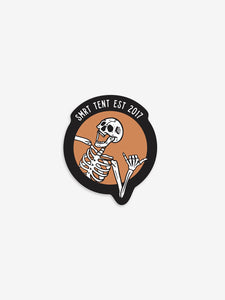 Pictured is a laughing white skeleton in-frame from the ribs up. All the fingers of the left hand are bent except for the thumb and pinky finger. The other hand and arm are out of frame. The backdrop is orange and outlined thickly with another frame in black where it says SMRT Tent Est 2017 in white text. Part of the left arm breaks out of the orange circle frame and overlaps with the black frame. Perfect to put on your car's dash or roof top tent! Available for sale by SMRT Tent in Denver, Colorado, USA.