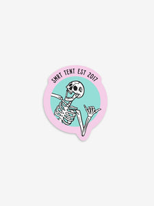 Pictured is a laughing white skeleton in-frame from the ribs up. All fingers of the left hand are bent except for the thumb and pinky finger. The other hand and arm are out of frame. The backdrop is teal and outlined thickly with another frame in pink where it says SMRT Tent Est 2017 in black text. Part of the left arm breaks out of the teal circle frame and overlaps with the pink frame. Perfect to put on your truck's dash or roof top tent! Available for sale by SMRT Tent in Denver, Colorado, USA.