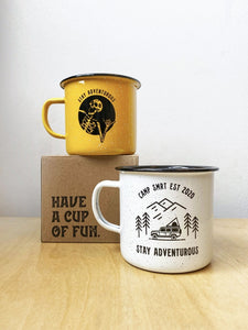 Pictured are the two variants of the SMRT Tent enamel mug. One is the Stay Adventurous Skeleton mug in yellow, set atop a brown cardboard box that says Have A Cup Of Fun. On the table and slightly to the right is the Camp SMRT EST 2020 Stay Adventurous in white. Available for purchase by SMRT Tent in Denver, Colorado, USA.
